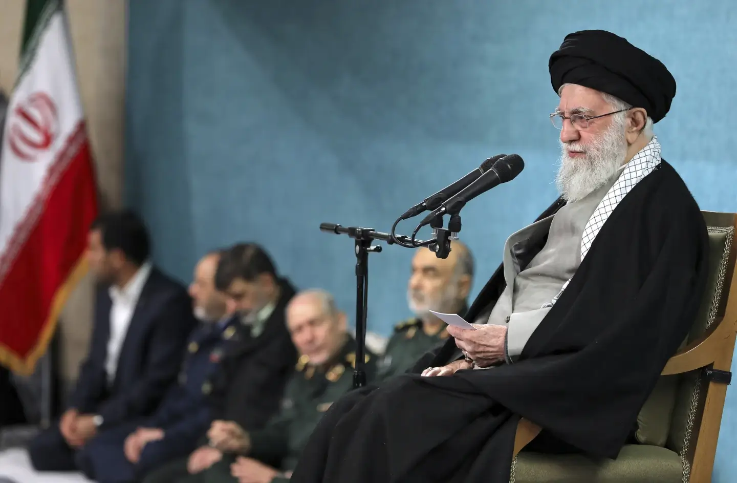 Ayatollah Ali Khamenei, seated with a microphone, addresses Iranian generals in a room with an Iranian flag and Khomeini portrait, 2025.